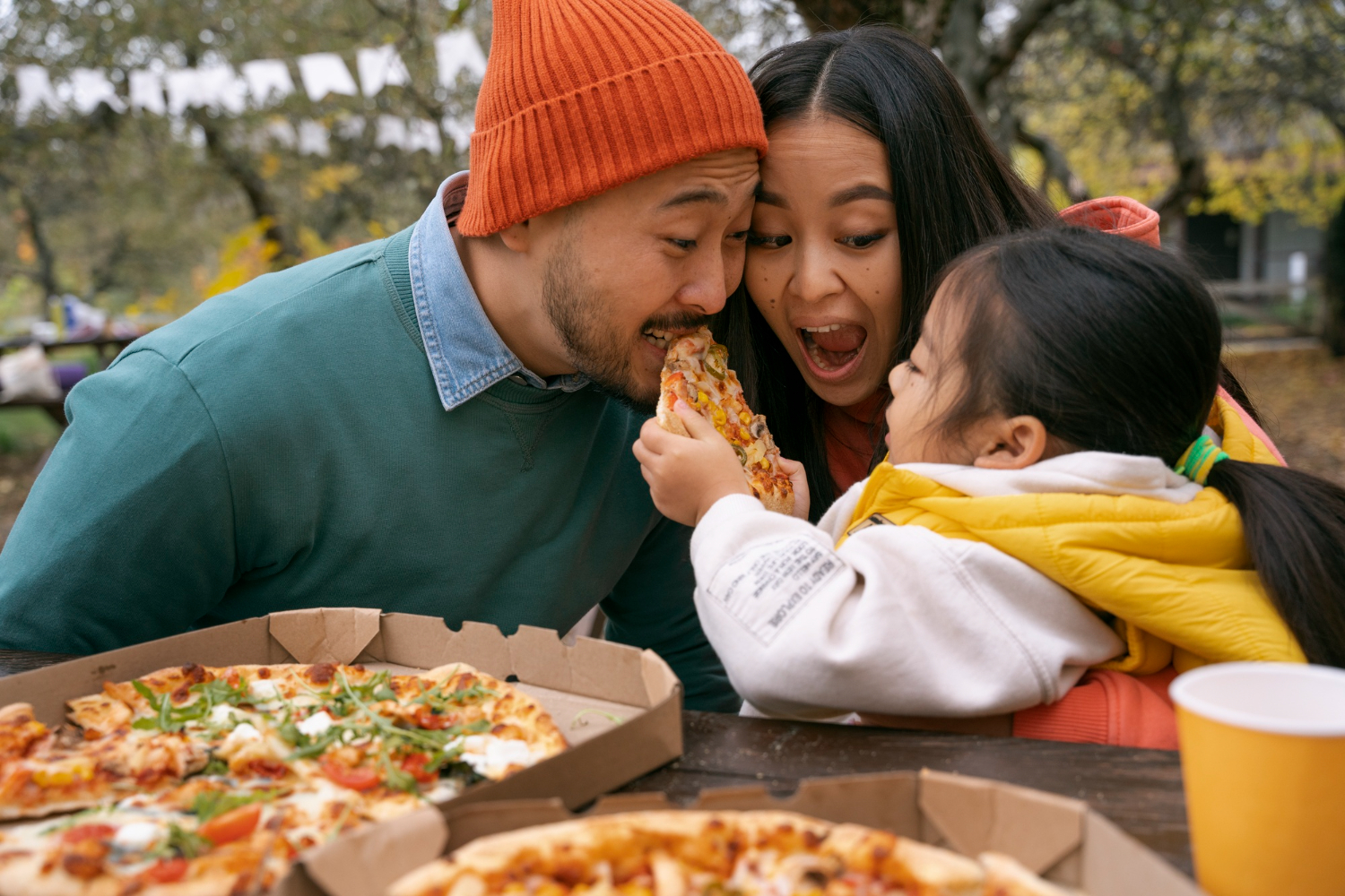 un papa et une maman qui mange une pizza avec leur fille dans un parc